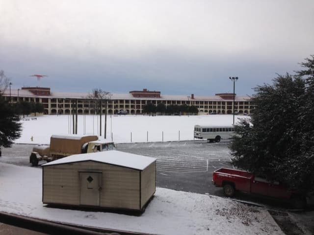 A rare snowfall covers the training grounds at Fort Benning, with a parked bus and vehicles under an overcast sky