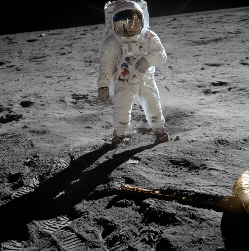 Astronaut Buzz Aldrin standing on the lunar surface during the Apollo 11 mission, his white spacesuit lit by the sun against the dark sky, footprints visible across the gray regolith and the reflection of fellow astronaut Neil Armstrong in his visor