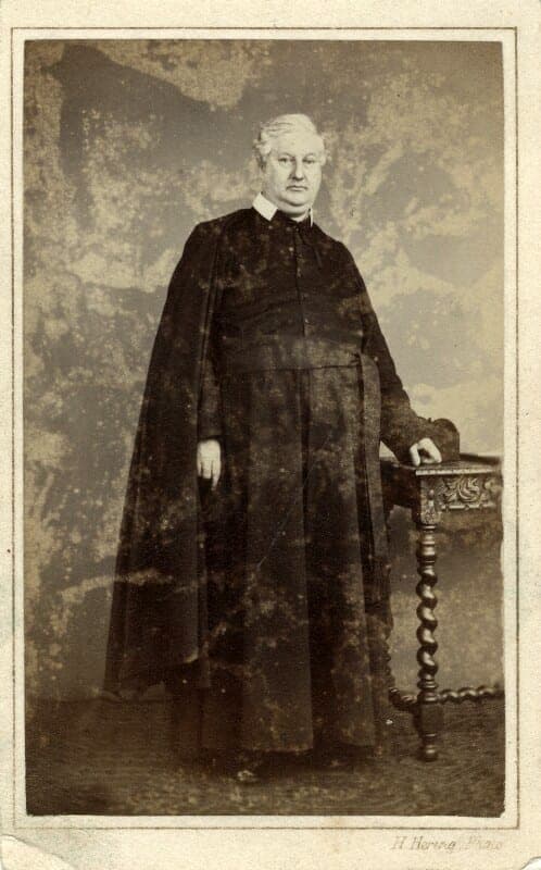 Frederick William Faber, the Anglican convert to Catholicism who wrote the hymn 'Faith of Our Fathers', photographed standing in a long clerical cassock and cloak beside a carved side table