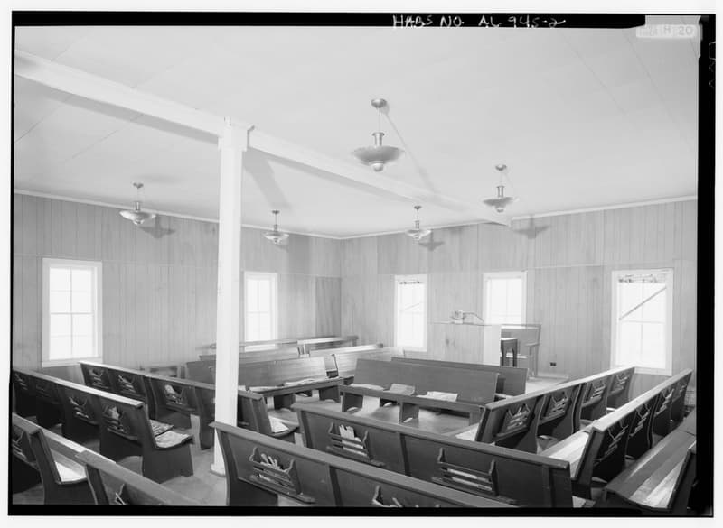The interior of a small wooden country Baptist church photographed from the back, looking obliquely across rows of plain pews toward a raised pulpit, with wire racks fixed to the pew backs holding hymnals and paper hand fans