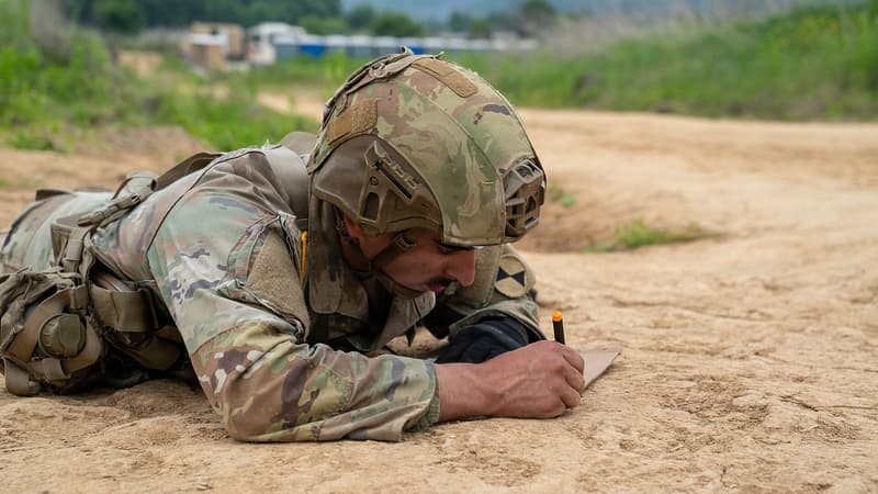 A soldier lies prone on a dirt trail plotting a point on his map during Army land navigation training.