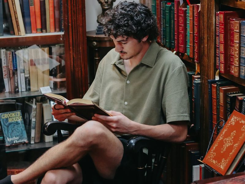 A man reads a book alone in a dimly lit library, surrounded by shelves of old volumes