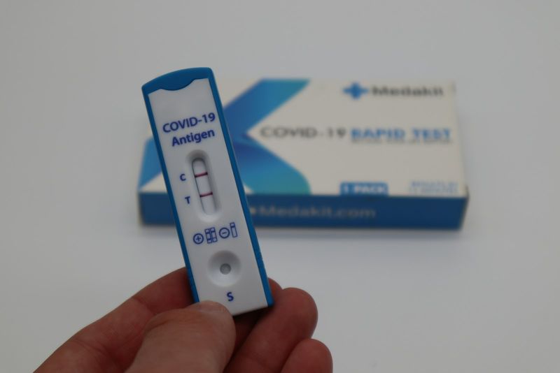 A hand holding a COVID-19 antigen rapid test cassette in front of its packaging