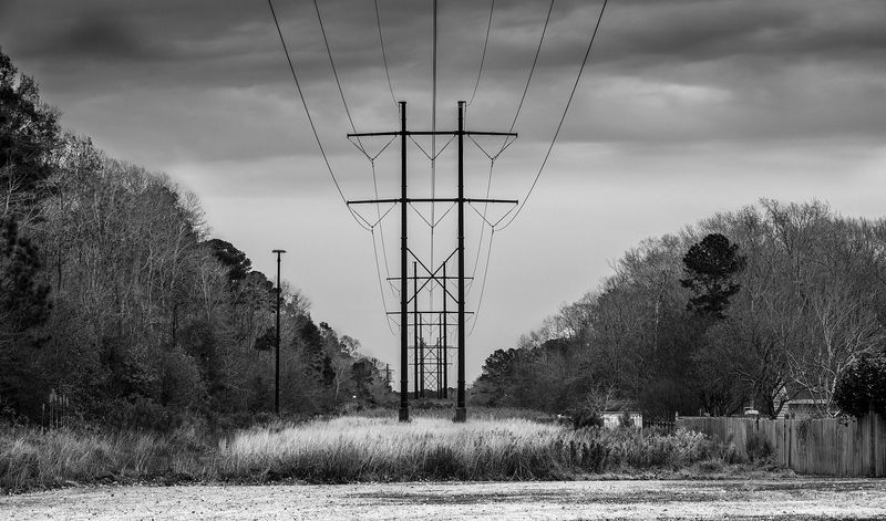Black-and-white photograph of a high-voltage transmission tower marching across a rural clearing at dusk — the kind of utility line easement the post discusses opposing through the APSC and (failing that) the eminent domain process.