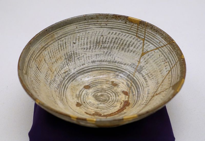 A beige Korean stoneware tea bowl whose cracks have been mended with bright gold lacquer in the kintsugi style, displayed on a purple cushion